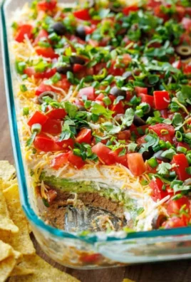 A colorful 7 layer dip featuring beans, cheese, guacamole, and toppings in a glass dish.