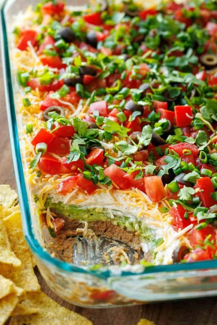 A colorful 7 layer dip featuring beans, cheese, guacamole, and toppings in a glass dish.